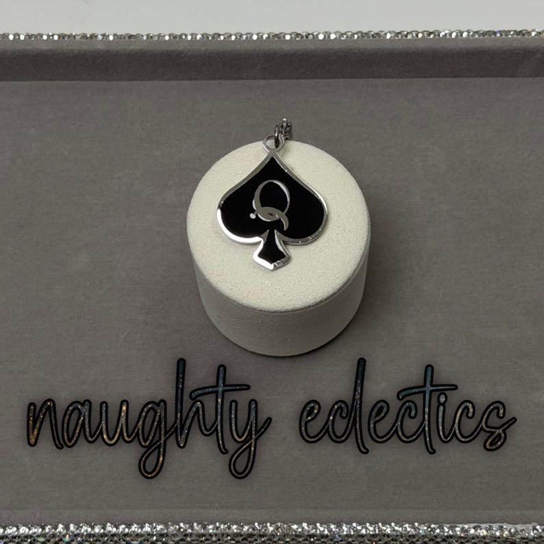 May include: A silver-toned pendant featuring a black spade design with a white 'Q' in the center. The pendant is displayed on a white cylindrical stand against a gray background. The words "naughty eclectics" are written in cursive below.