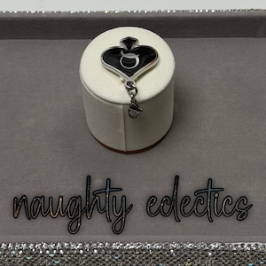 May include: A black and silver charm with a spade design, featuring a stylized letter "Q" in the center. The charm is attached to a small silver clasp. The words "naughty eclectics" are written in cursive.