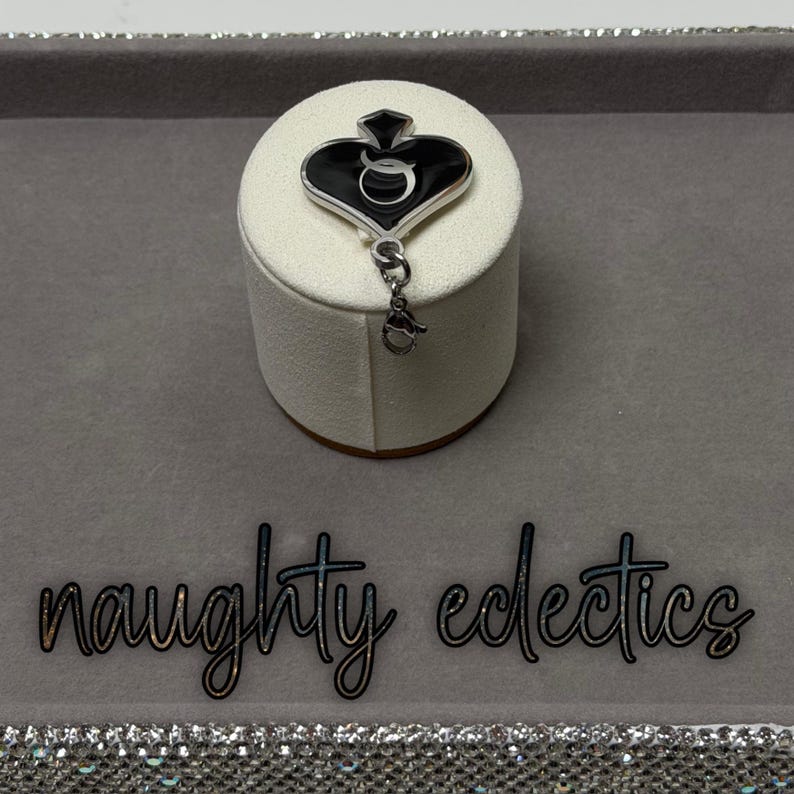 May include: A black and silver charm with a spade design, featuring a stylized letter "Q" in the center. The charm is attached to a small silver clasp. The words "naughty eclectics" are written in cursive.