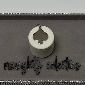 May include: A silver-colored spade pendant necklace. The pendant is displayed on a white cylindrical stand, set against a gray background. The words "naughty eclectics" are written in cursive.