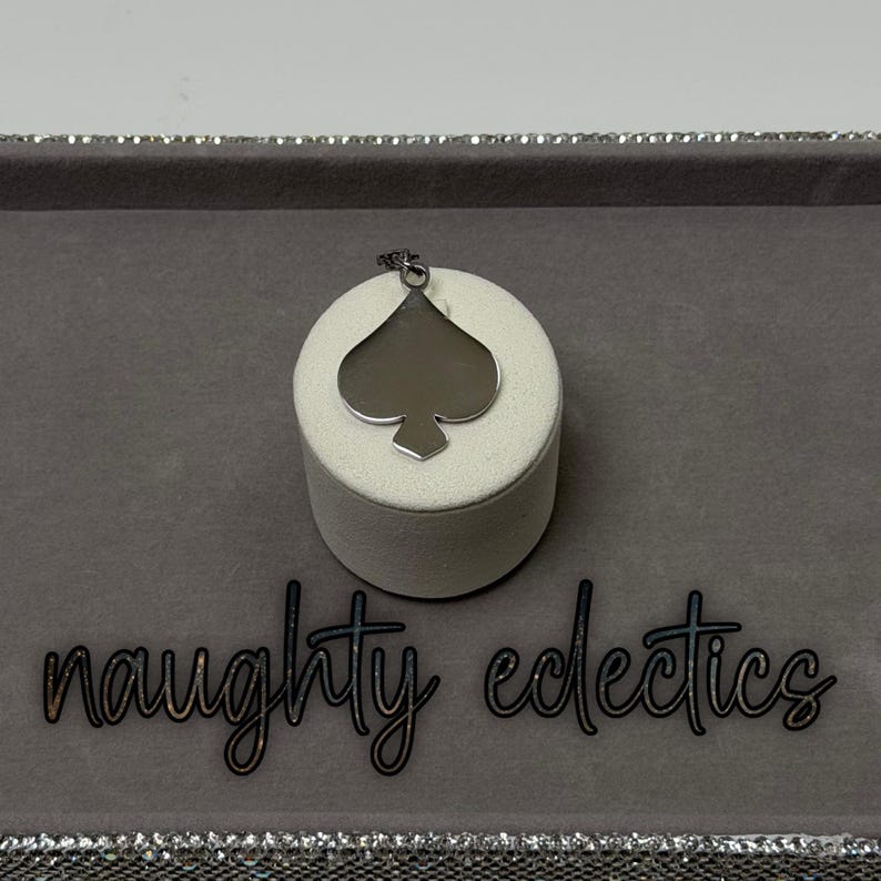 May include: A silver-colored spade pendant necklace. The pendant is displayed on a white cylindrical stand, set against a gray background. The words "naughty eclectics" are written in cursive.