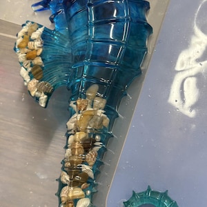 Könnte beinhalten: Eine durchscheinende blaue Seepferdchen-Skulptur mit einem hohlen Körper, der mit verschiedenen kleinen Muscheln und Kieselsteinen gefüllt ist. Das Seepferdchen hat ein detailliertes Auge, einen geschwungenen Schwanz und eine Seitenflosse mit Muscheln.