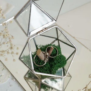 May include: Geometric glass ring box with silver trim, open to reveal two rings on a bed of green moss. The box is a clear, multi-faceted shape, reflecting light. A rustic wedding theme.