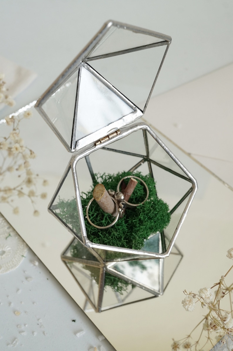 May include: Geometric glass ring box with silver trim, open to reveal two rings on a bed of green moss. The box is a clear, multi-faceted shape, reflecting light. A rustic wedding theme.