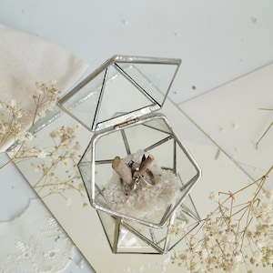 May include: A geometric glass ring box with an open lid, showcasing two rings nestled on white crystals. The box is set on a mirrored surface, with dried flowers adding a delicate touch. The box is approximately 3 inches wide.