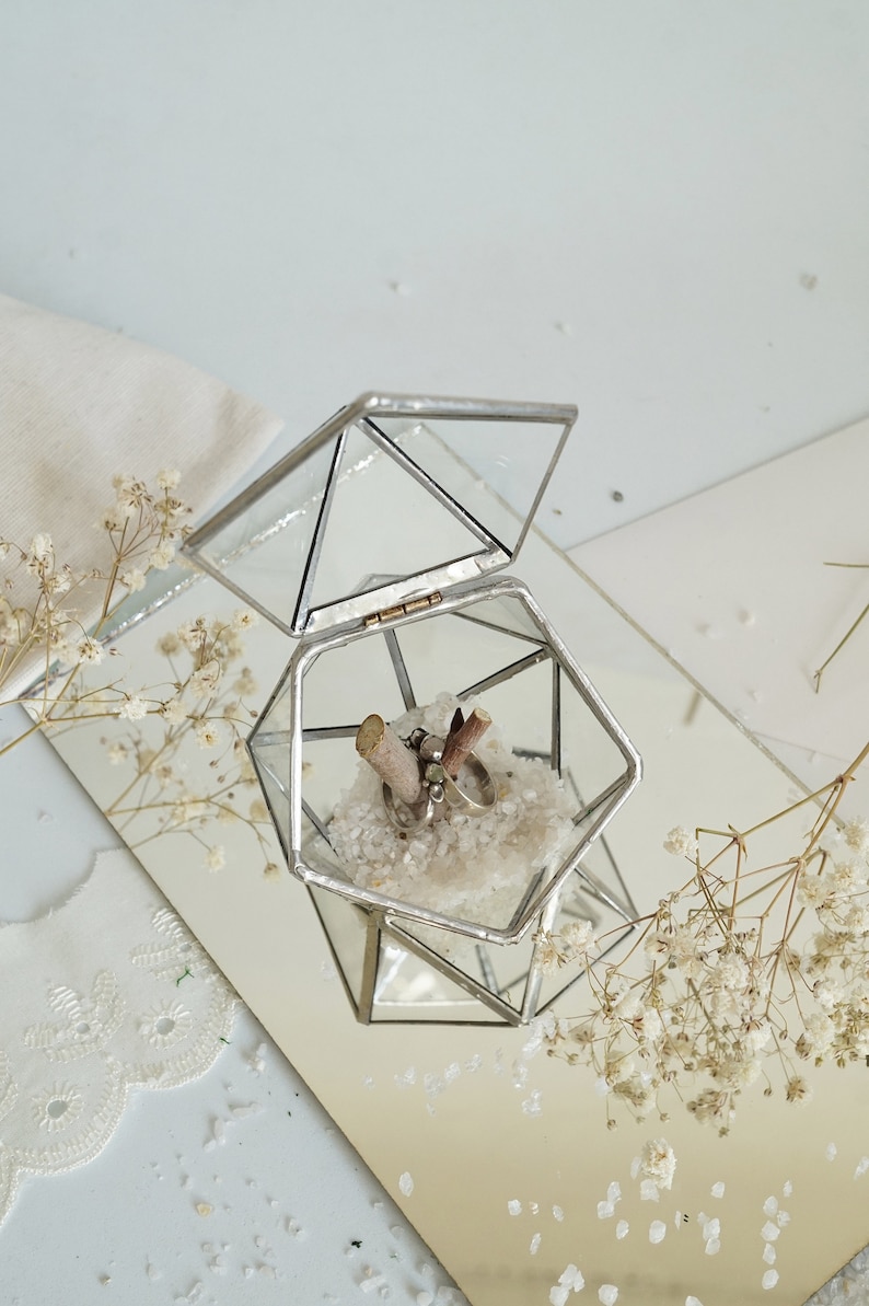 May include: A geometric glass ring box with an open lid, showcasing two rings nestled on white crystals. The box is set on a mirrored surface, with dried flowers adding a delicate touch. The box is approximately 3 inches wide.