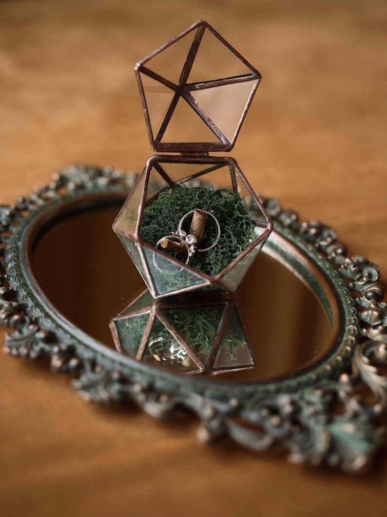 May include: A geometric glass ring box with a copper frame, open to reveal two rings nestled in green moss. The box sits on a decorative, antique-style mirror, reflecting the box and rings. The background is a warm, neutral tone.