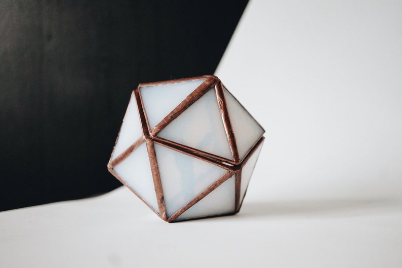 May include: Geometric glass terrarium with a copper-colored metal frame. The terrarium is a multi-faceted shape with white glass panels. The background is split between black and white. The object is likely used for displaying small plants or decorative items.