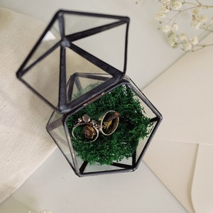May include: A geometric glass terrarium with a hinged lid, containing green moss and two rings. The rings are silver with decorative elements. The terrarium sits on a white surface, with a white envelope and dried flowers in the background.