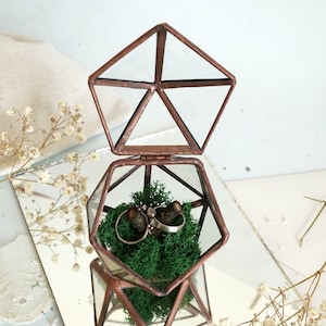 May include: A geometric glass and copper ring box, open to reveal two rings nestled in green moss. The box is pentagon-shaped with a hinged lid, reflecting on a mirrored surface. Delicate white flowers and a neutral fabric are in the background.
