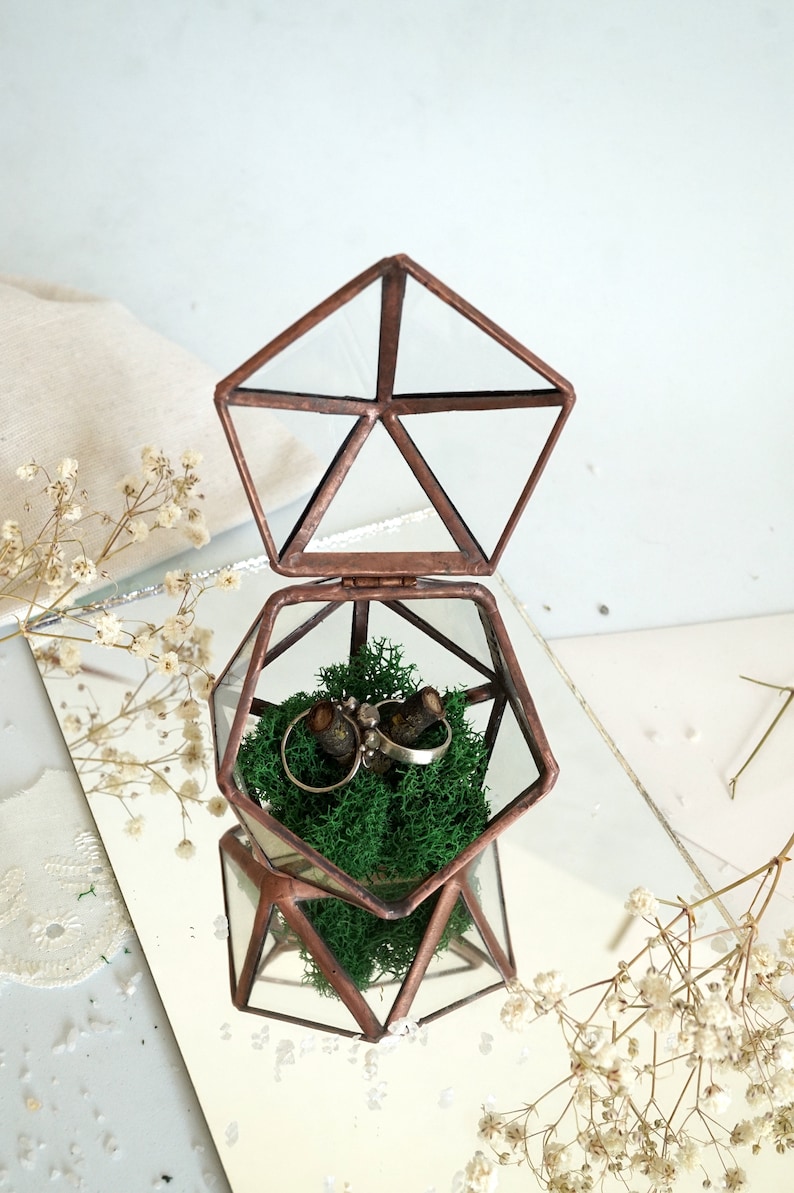 May include: A geometric glass and copper ring box, open to reveal two rings nestled in green moss. The box is pentagon-shaped with a hinged lid, reflecting on a mirrored surface. Delicate white flowers and a neutral fabric are in the background.