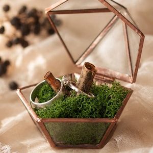 Op de afbeelding: Een geometrische glazen en koperen ringendoos, open om twee zilveren ringen en een stuk hout in groen mos te tonen. De doos staat op een crèmekleurige stof, wat een zachte, natuurlijke uitstraling creëert.