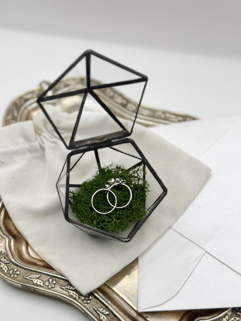May include: A geometric glass ring box with a hinged lid, containing two silver rings nestled in green moss. The box sits on a silver tray with a white envelope and a small white cloth bag.