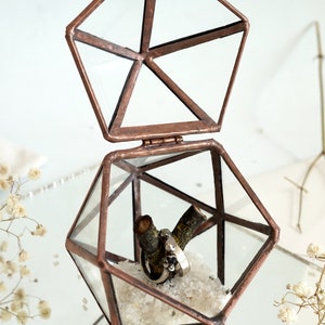 May include: A geometric terrarium with a hinged lid, crafted from copper-toned metal and clear glass. Inside, two rings rest on a small branch, surrounded by white crystals. The terrarium is open, revealing its contents.