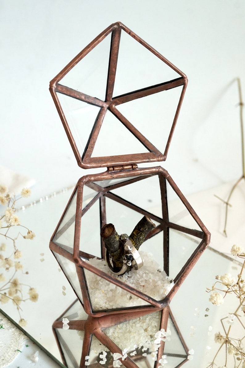 May include: A geometric terrarium with a hinged lid, crafted from copper-toned metal and clear glass. Inside, two rings rest on a small branch, surrounded by white crystals. The terrarium is open, revealing its contents.