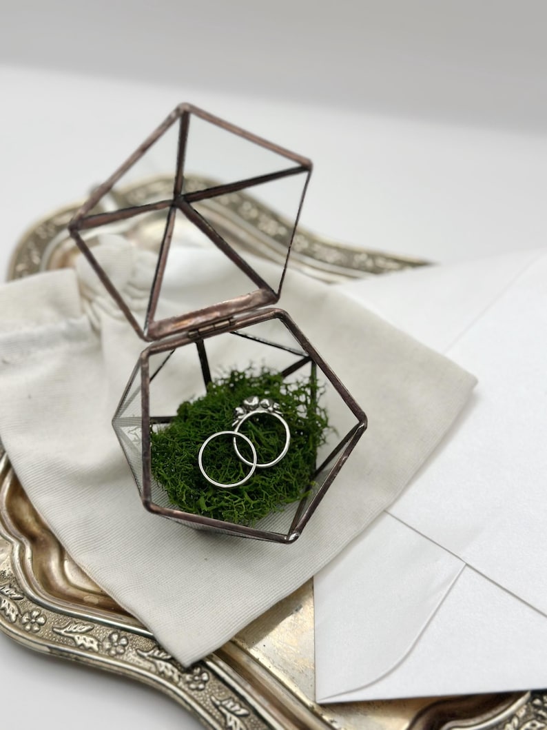 May include: A geometric glass ring box with a copper-colored frame, open to reveal two silver rings nestled in green moss. The box sits on a silver tray with a white envelope and a small cloth bag.