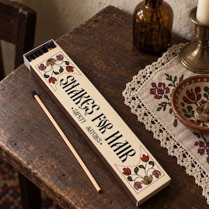 May include: A rectangular box of safety matches with a cream-colored label featuring floral illustrations in red, purple, and green. The text on the box reads "SHAKES FOR HAIR" and "SAFETY MATCHES." A single long matchstick with a black tip lies beside the box.