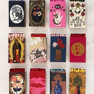 May include: Twelve rectangular matchboxes arranged in a grid, each featuring unique graphic illustrations including floral patterns, a hand with an eye, a portrait, lips, and text reading SHAKE FOR HAIR, with various colored matchstick tips visible.