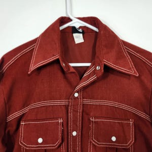 May include: A rust-colored button-up shirt with white stitching and two chest pockets. The shirt has a pointed collar and is hanging on a white hanger. The stitching is visible on the collar, pockets, and seams.