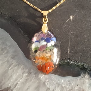 Op de afbeelding: Goudkleurige ketting met een ovale hanger. De hanger is van heldere hars en bevat lagen van kleurrijke edelstenen: rood, oranje, geel, groen, blauw en paars. De ketting is geplaatst tegen een donkere achtergrond.