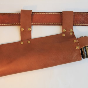 May include: A brown leather sheath with brass rivets and a belt loop. The sheath is designed to hold a knife or other tool.