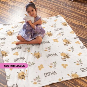 May include: A white fleece blanket with a repeating pattern of cartoon bees, honeycomb shapes, and floral accents. The name "SOPHIA" is printed throughout the design. The blanket is customizable, as indicated by the pink text in the lower left corner.