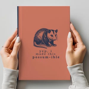 Peut inclure: Un carnet de couleur corail avec une tranche bleu marine. La couverture présente une illustration détaillée d'un opossum et le texte "yep, i made this possum-ible" en bleu foncé. Le carnet est tenu par deux mains.