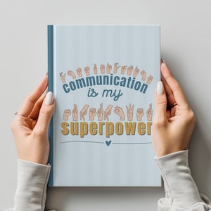 以下が含まれることがあります： 縦縞模様のライトブルーのノート。ダークブルーとゴールドで「communication is my superpower」という文字が書かれています。手話の指文字を表す手のイラストと、小さなハートのアイコンがデザインされています。