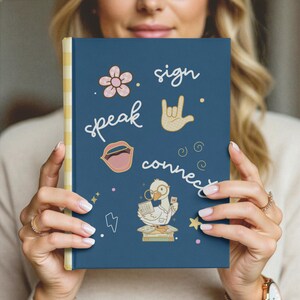 May include: A dark blue hardcover journal with a yellow and white striped spine. The cover features a pink flower, a hand sign, an open mouth, and a cartoon duck wearing glasses reading a book. The text reads sign, speak, and connect.