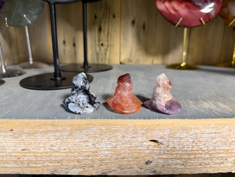 May include: Three small, carved stone figurines shaped like pointed hats. The first is speckled black and white, the second is a translucent reddish-orange, and the third is a pale, semi-translucent pinkish-purple with a lighter base.