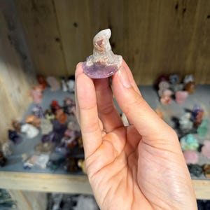 May include: A small, carved crystal figurine shaped like a pointed hat. The object features a smooth, rounded base and a tapered top, displaying a mix of translucent white, pale purple, and earthy brown mineral inclusions throughout the stone structure.
