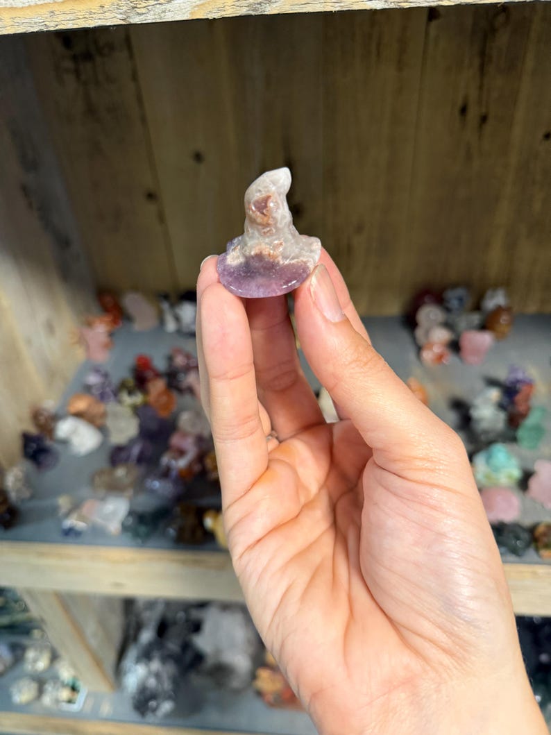 May include: A small, carved crystal figurine shaped like a pointed hat. The object features a smooth, rounded base and a tapered top, displaying a mix of translucent white, pale purple, and earthy brown mineral inclusions throughout the stone structure.