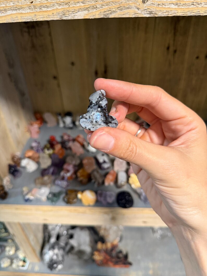 May include: A small, polished stone carving in the shape of a seated figure. The stone features a mottled pattern of black, white, and gray with a distinct iridescent blue flash visible on the front surface, showcasing a smooth, rounded texture.