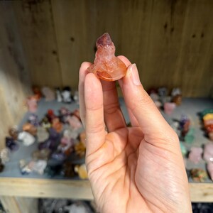 May include: A small, translucent, reddish-orange carved crystal figurine shaped like a seated figure. The stone features natural variations in color, ranging from deep burnt orange to lighter, clearer sections, with a smooth, polished surface texture.