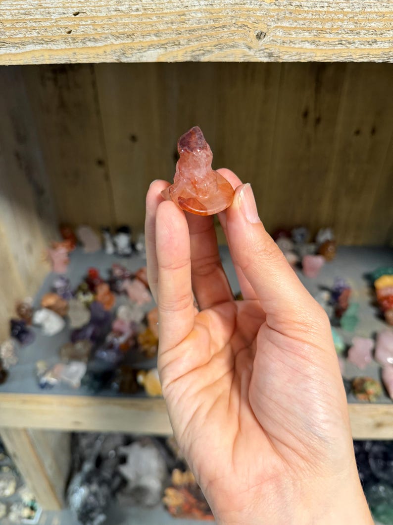 May include: A small, translucent, reddish-orange carved crystal figurine shaped like a seated figure. The stone features natural variations in color, ranging from deep burnt orange to lighter, clearer sections, with a smooth, polished surface texture.