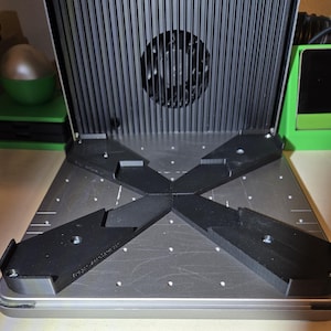 May include: A close-up of a silver 3D printer bed with four black plastic corner supports. The supports are designed to hold a print in place during the printing process.