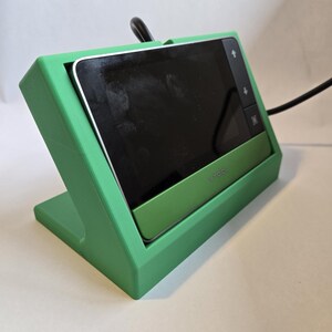 May include: A green plastic stand with a black and silver device inside. The device has a screen and buttons. The stand has a cutout for the device's power cord.