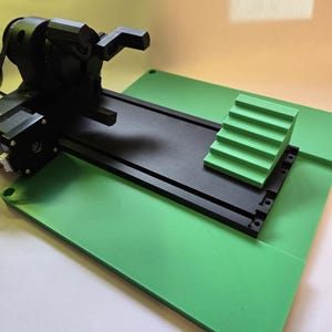May include: A black robotic arm with a green platform and a green staircase-shaped object. The arm is mounted on a black track.