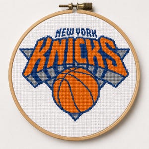 May include: A circular wooden embroidery hoop containing white fabric with a cross-stitch design of the New York Knicks basketball team logo. The design features the orange and blue text NEW YORK KNICKS above an orange basketball with blue outlines.