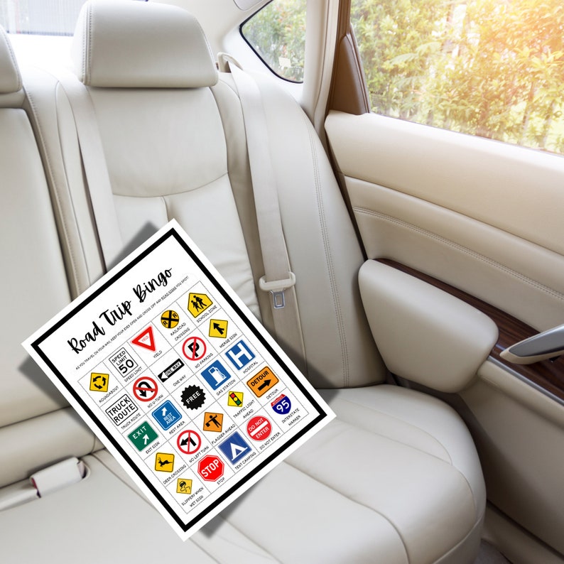 May include: A white sheet of paper with a black border featuring "Road Trip Bingo" text and various road sign illustrations. The bingo card is set against a beige car seat.
