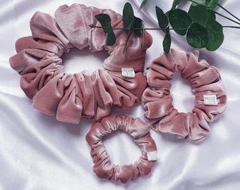 May include: Four dusty rose velvet scrunchies of varying sizes are arranged on a white satin surface. The scrunchies have a soft, gathered texture. A sprig of green leaves is placed near the scrunchies. Each scrunchie has a small, white tag.