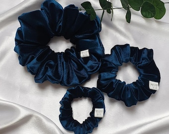May include: Four navy blue velvet scrunchies of varying sizes are arranged on a white satin surface. Each scrunchie has a small white tag. Green leaves are in the background.