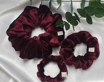 May include: Four burgundy velvet scrunchies of varying sizes are arranged on a white satin surface. Each scrunchie has a small, square white tag with a butterfly logo. Green leaves are visible in the background.