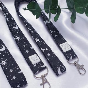 May include: Black lanyards patterned with white stars and crescent moons. Each lanyard features a silver-coloured metal clasp and a small white tag with a butterfly design. The lanyards are shown on a white satin surface.