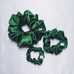 May include: Three green satin scrunchies of different sizes. The largest scrunchie is in the centre, with a smaller scrunchie to the right and the smallest scrunchie below the centre scrunchie.