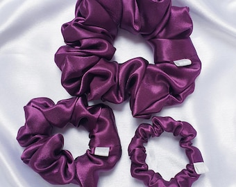 May include: Three purple satin scrunchies of different sizes. The largest scrunchie is in the center, with a smaller scrunchie to the left and the smallest scrunchie to the right.