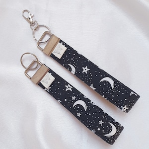 May include: Two black and white keychains with silver key rings. The keychains feature a moon and star pattern with a white butterfly design on the strap.