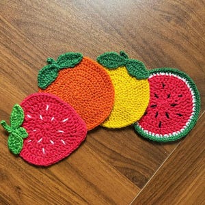 May include: A collection of handmade crochet coasters in the shape of fruits. The set includes a red strawberry, an orange, a yellow lemon, and a red watermelon. Each coaster has green leaves and is made of yarn.