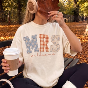 May include: An off-white short-sleeve t-shirt featuring the text MRS in large block letters with a blue floral, orange gingham, and beige floral pattern, with the word williams in cursive script below, paired with black leggings and cream leg warmers.