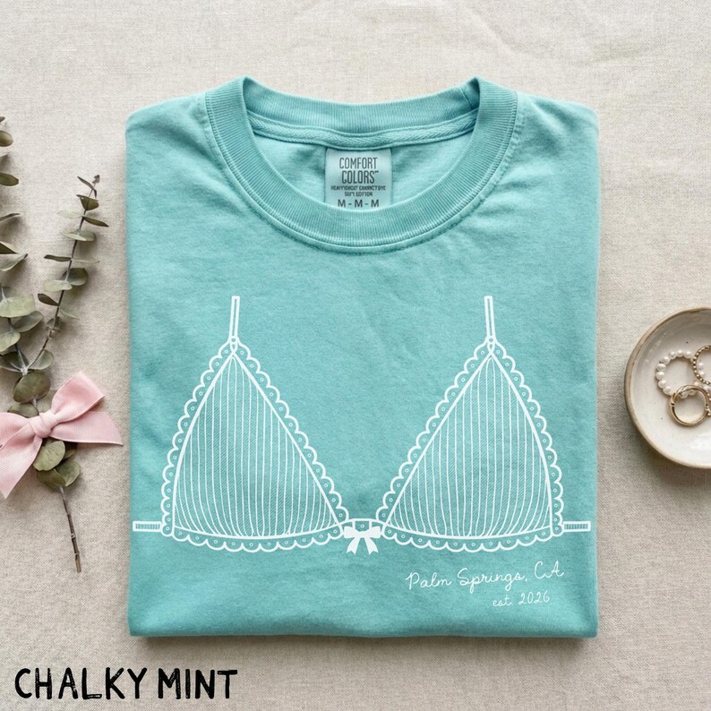 Puede incluir: Una camiseta de color menta con un gr&aacute;fico blanco de un sujetador. La camiseta tiene cuello redondo y mangas cortas. El texto "Palm Springs, CA est 2026" est&aacute; impreso en la camiseta.
